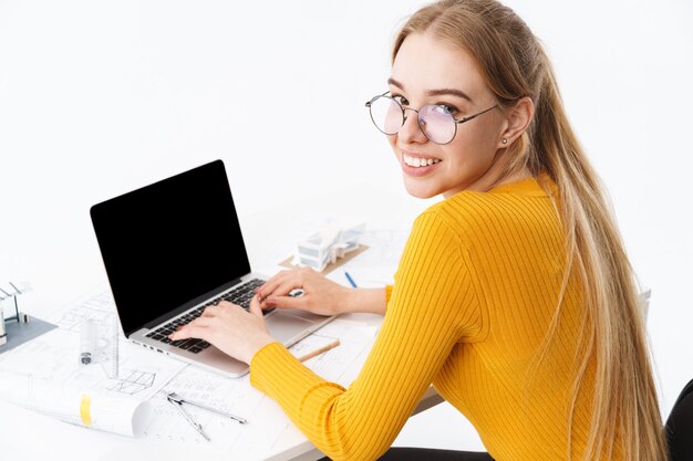 Side view of a beautiful young woman sitting at the workplace working on blank screen laptop computer isolated over white wall