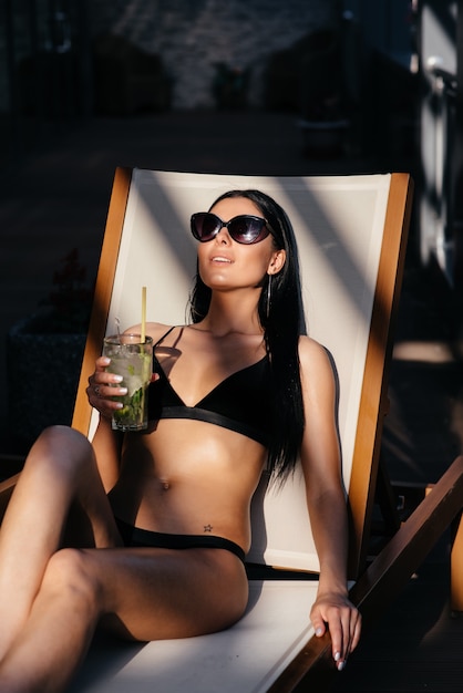 Side view of beautiful young woman in sunglasses and swimsuit holds a glass with cocktail, looking at the sea, dreaming, while resting on luxury sunbed
