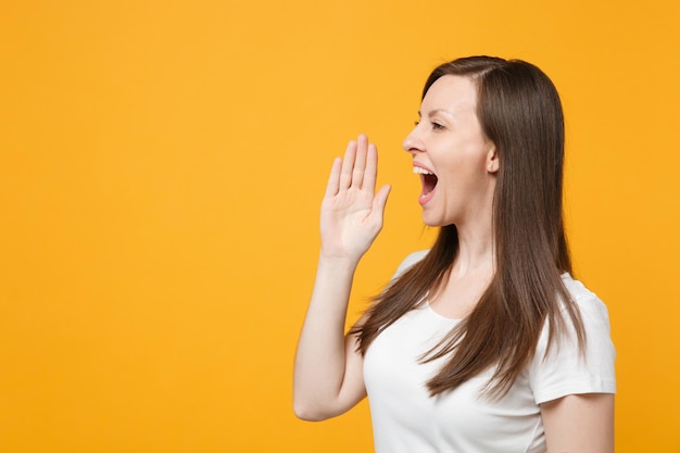 Side view of beautiful young woman in white casual clothes looking aside, screaming with hand gesture isolated on yellow orange wall background in studio. people lifestyle concept. mock up copy space