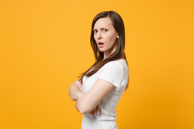 Side view of bewildered young woman in white casual clothes looking camera, holding hands crossed isolated on bright yellow orange background in studio. People lifestyle concept. Mock up copy space.