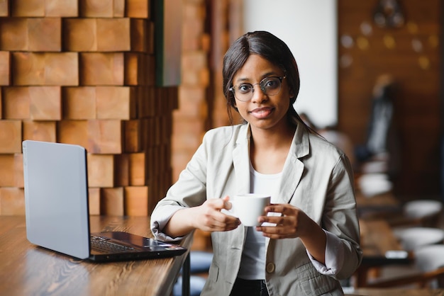 Вид сбоку на чернокожую даму, наслаждающуюся утренним кофе и проверяющую электронную почту на пустом пространстве внутри кафе с ноутбуком