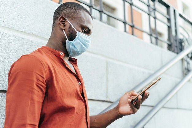 Side view of black man wearing surgical mask and orange shirt. he is is using smartphone in the street