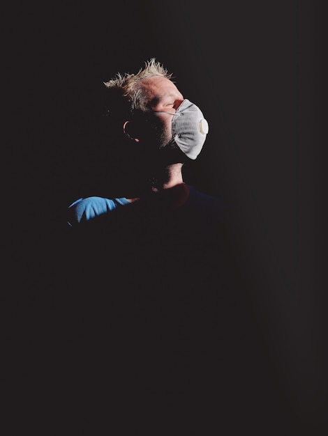 Side view of blond haired male shaper in protective respirator mask and with closed eyes in darkness against black background illuminated by sunlight