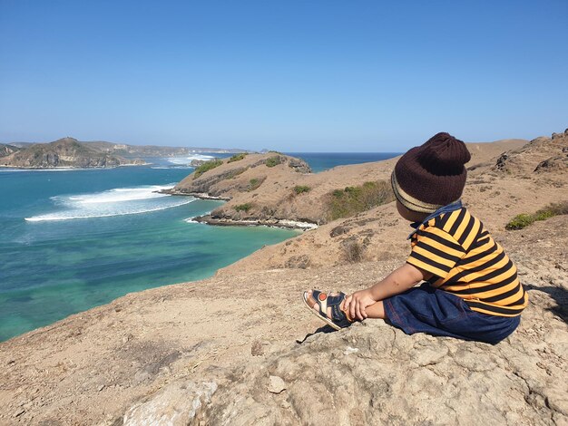 Side view of boy sitting on rock by sea against clear sky