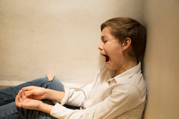 Foto vista laterale di un ragazzo che sbadigli mentre è seduto contro il muro