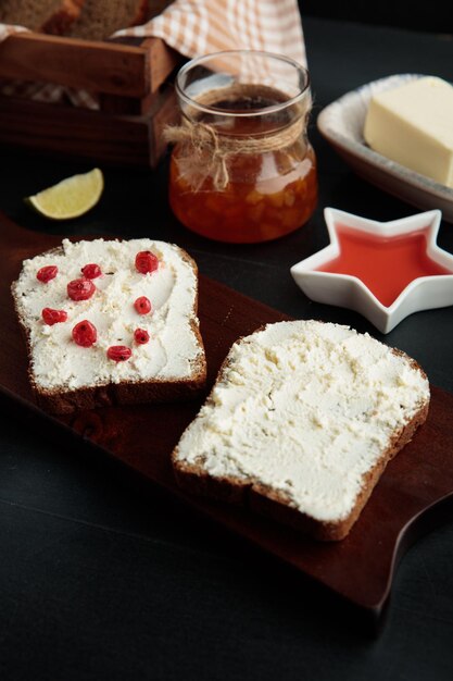 Side view of bread slices smeared with cottage cheese with red
currants on it on cutting board with jam butter lime slice bread
slices on black background