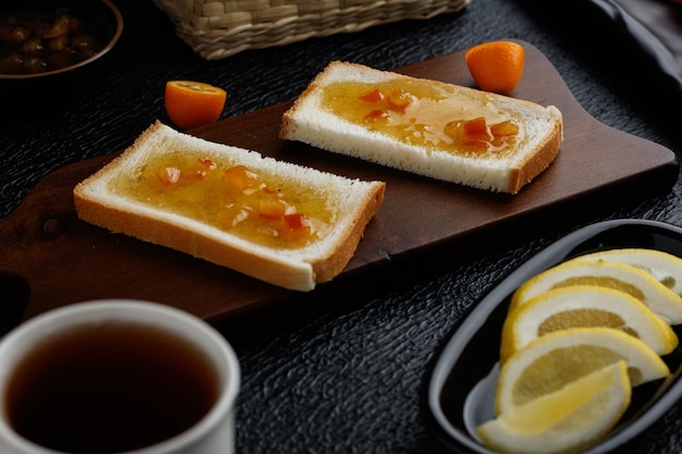 まな板のパンのスライスにジャムを塗った朝食セットと黒の背景にハーフカットのヒメタチバナとお茶のレモンスライスの側面図