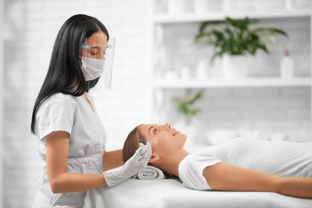 Side view of brunette beautician in protective mask doing beauty injection for improvements skin face