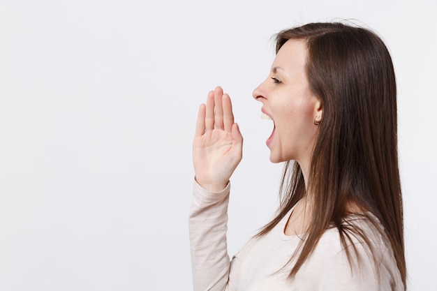 Side view of brunette young woman in light clothes screaming with hand gesture near mouth isolated on white wall background in studio. People sincere emotions lifestyle concept. Mock up copy space.