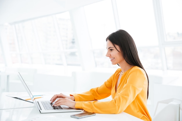 Vista laterale della donna d'affari in camicia arancione che usa il computer portatile e si siede al tavolo in ufficio