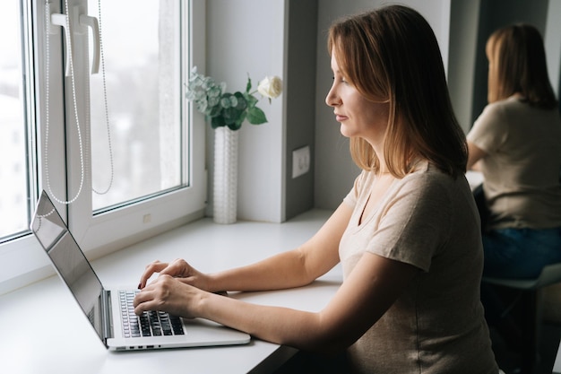 Side view of business woman typing on laptop keyboard sitting at window sill working from home office at cozy atmosphere