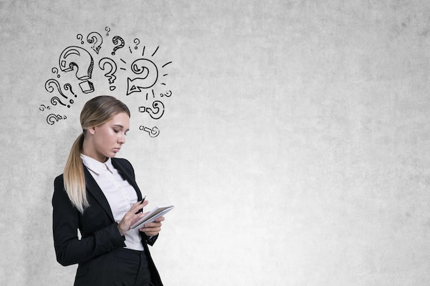 Side view of a businesswoman with a notebook writing near a concrete wall with many question marks drawn on it. Mock up