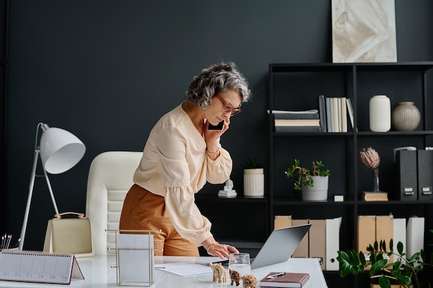 Side view of busy mature female boss with smartphone by ear bending over desk