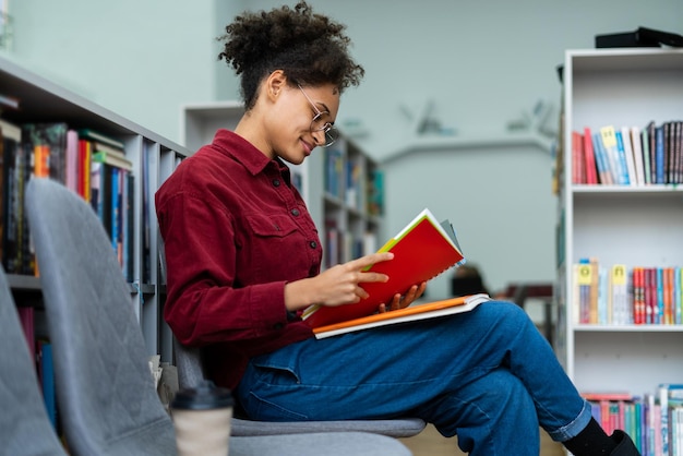 Photo side view of the calm multiracial lady sitting at the chair and reading book with pleasure smile studying concept