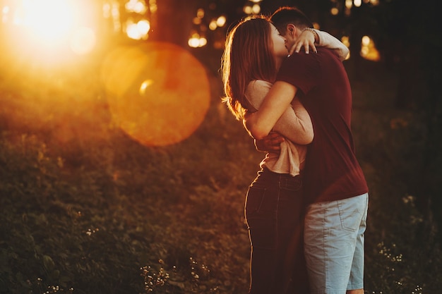 Side view of casual loving couple embracing while standing in green park with glowing sunset light