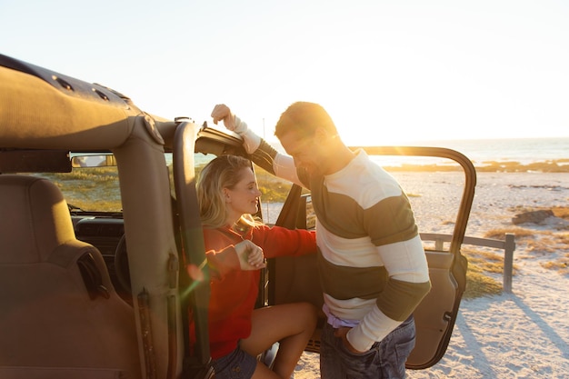 Side view of a Caucasian couple inside an open top car with its door open, looking at each other, smiling and holding hands