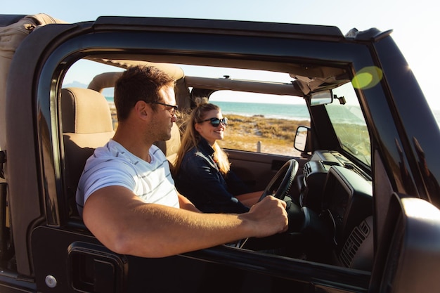 Side view of a Caucasian couple sitting inside an open top car with blue sky and sea in the background, driving and smiling