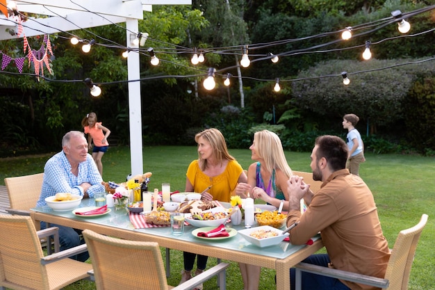 Side view of a Caucasian multi-generation family in the garden, the adults sitting at a table for a meal together and talking, while the granddaughter and grandson play in the background