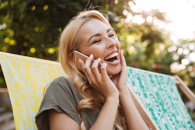 サンラウンジャーに座って、屋外で電話で話しているドレスを着た陽気なブロンドの女性の側面図