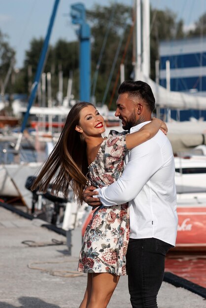 Side view of cheerful couple in love hugging in harbor with boats on sunny day in summer