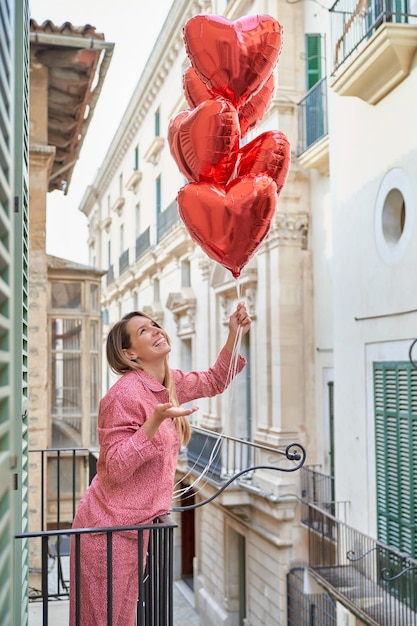 Foto vista laterale di una donna allegra in pigiama che guarda un mazzo di palloncini a forma di cuore mentre si trova sul balcone dell'hotel