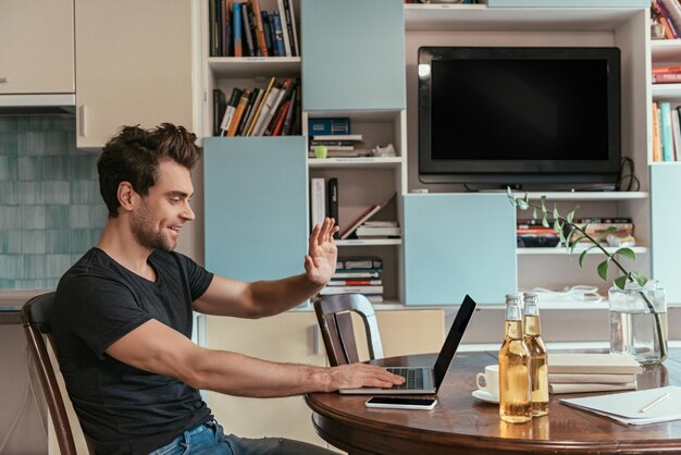 Photo side view of cheerful man waving hand during video chat near bottles of beer