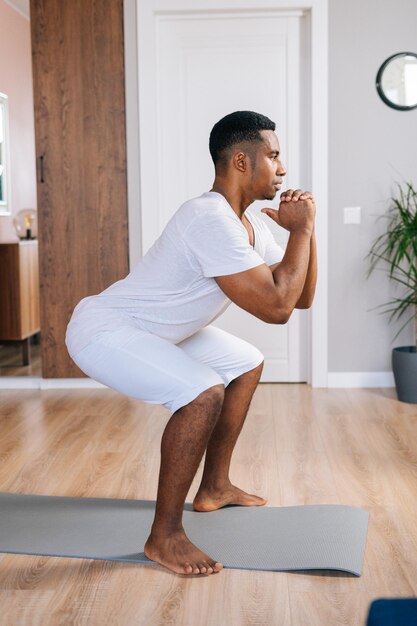 Vista laterale dell'uomo afroamericano muscoloso allegro che si esercita e fa squat durante l'allenamento in una luminosa stanza domestica. concetto di allenamento sportivo in palestra a casa.