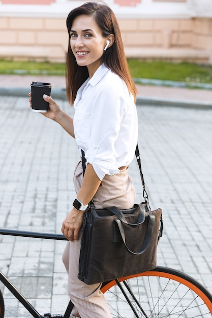 Вид сбоку веселой молодой женщины в летней одежде, гуляющей по городской улице с велосипедом и пьющей кофе на вынос