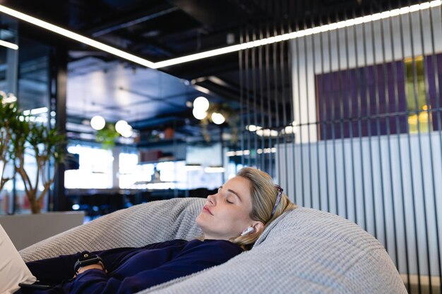 사진 스마트한 옷과 이어폰을 착용한 백인 사업가의 측면 뷰는 현대적인 사무실에서 일하는 동안 휴식을 취하고, 콩주머니에 누워 쉬고 있습니다.