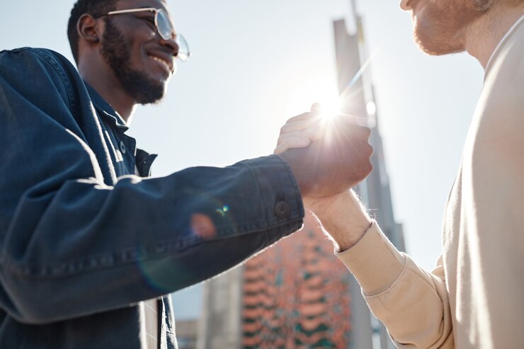 Photo side view close up of two men shaking hands while greeting each other outdoors in city lit by sunlight