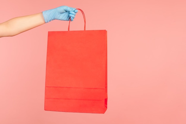 Side view closeup of human hand in blue surgical gloves holding and showing red shopping bag.