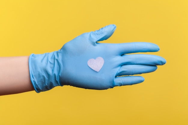 Side view closeup of human hand in blue surgical gloves holding small pink heart shape in hand