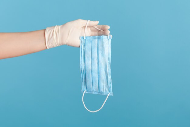 Side view closeup of human hand in white surgical gloves holding blue surgical medical mask.