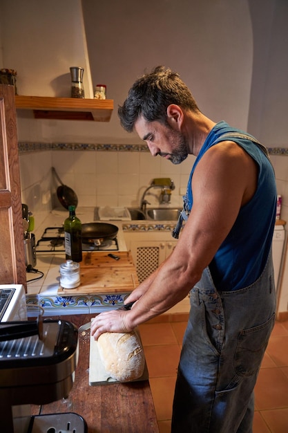 Side view of concentrated bearded man in denim overalls standing near counter and cutting bread while preparing breakfast in kitchen at morning