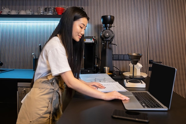 集中したカフェの従業員がラップトップを使いながら開いた口座帳に触れる側面の景色