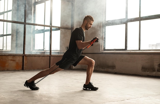 Photo side view of concentrated male athlete in sportswear doing lunge with elastic band on floor in gymnasium