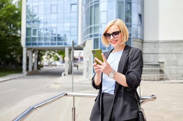 Side view of concentrated woman in stylish outfit text messaging on mobile phone while walking along near contemporary building