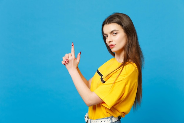 Side view of confident beautiful young woman in vivid casual clothes looking camera, holding hands like gun isolated on bright blue background in studio. People lifestyle concept. Mock up copy space.