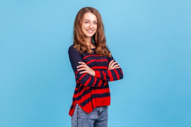 Side view of confident woman wearing striped casual style sweater standing with folded arms looking at camera with happy expression good mood Indoor studio shot isolated on blue background