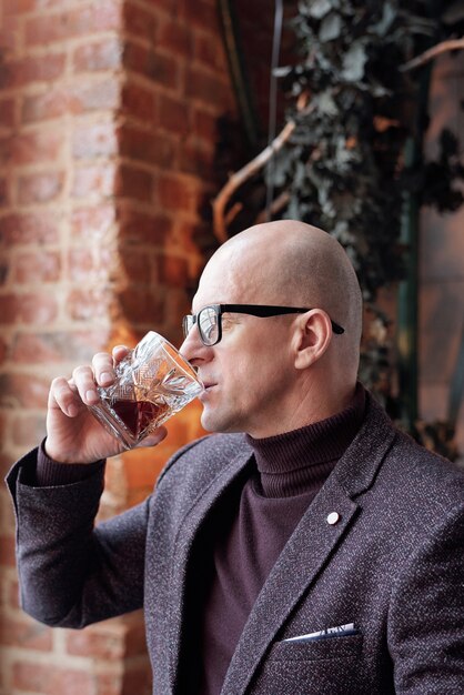 Side view of contemplative middle-aged businessman in glasses drinking whisky in loft bar