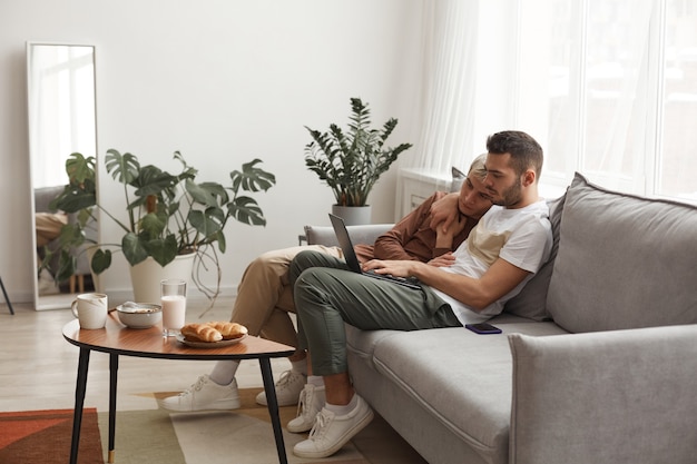 Side view at contemporary gay couple in everyday life, two young men using laptop together while relaxing on couch at home, copy space