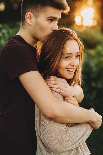 Side view of content young man and woman standing in bright sunset light and embracing with love in park
