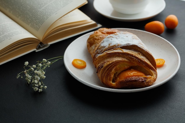 Side view of croissant with kumquat slices in plate and cup of tea open book with whole kumquats and little flower on black background