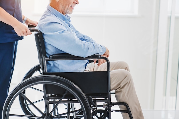 Обрезанный снимок медсестры, толкающей раненого пожилого пациента на инвалидной коляске в помещении, вид сбоку