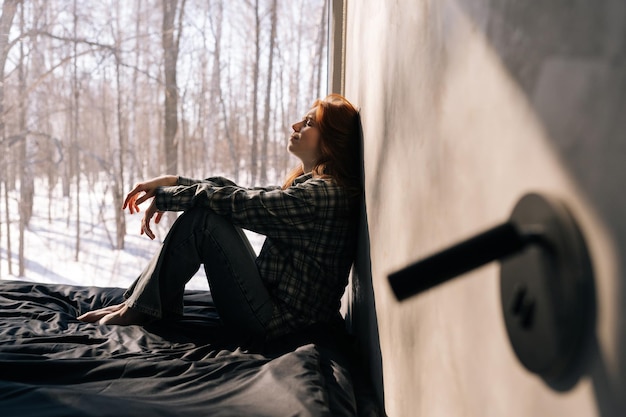 Side view of crying young woman sitting by window on sunny day on windowsill with winter nature