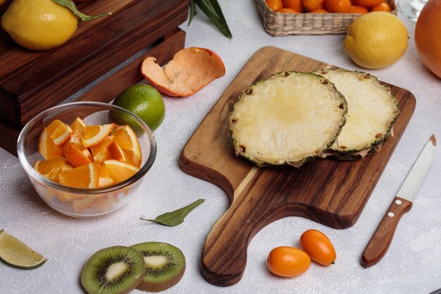 Side view of cut pineapple on cutting board with orange lemon kumquat with knife on white background