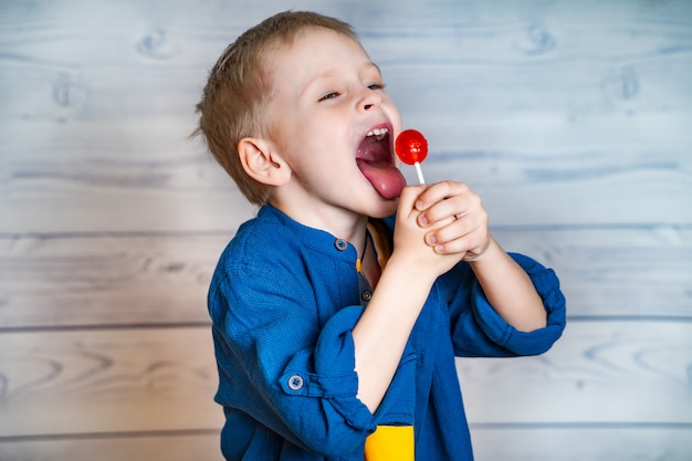La vista laterale di un bambino sveglio in camicia blu con la bocca ampiamente aperta sta mangiando una lecca-lecca rossa
