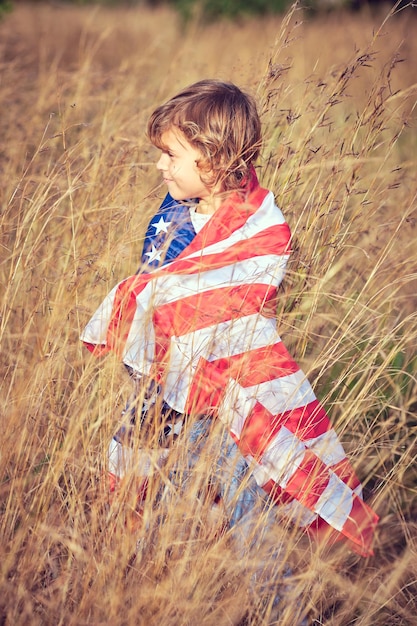 Side view of cute preteen boy wrapped in national flag of United States of America standing in rye meadow