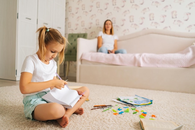 Side view of cute primary girl child writing homework in copybook sitting on floor with laptop for distance education online at home