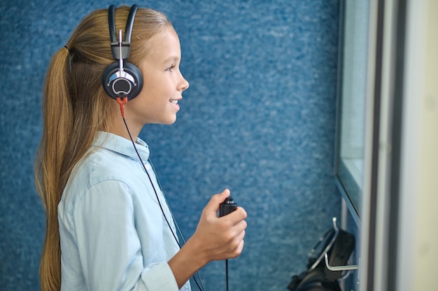 Side view of a cute young female patient in audiometer headsets standing in the soundproof booth